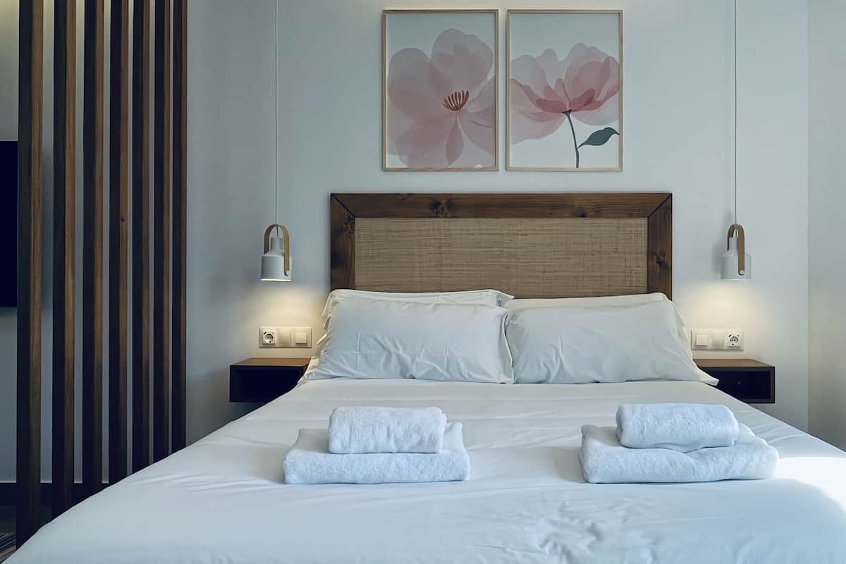 A neatly made bed with white linens and two folded towels, a wooden headboard, bedside lamps, and two framed floral paintings on the wall.