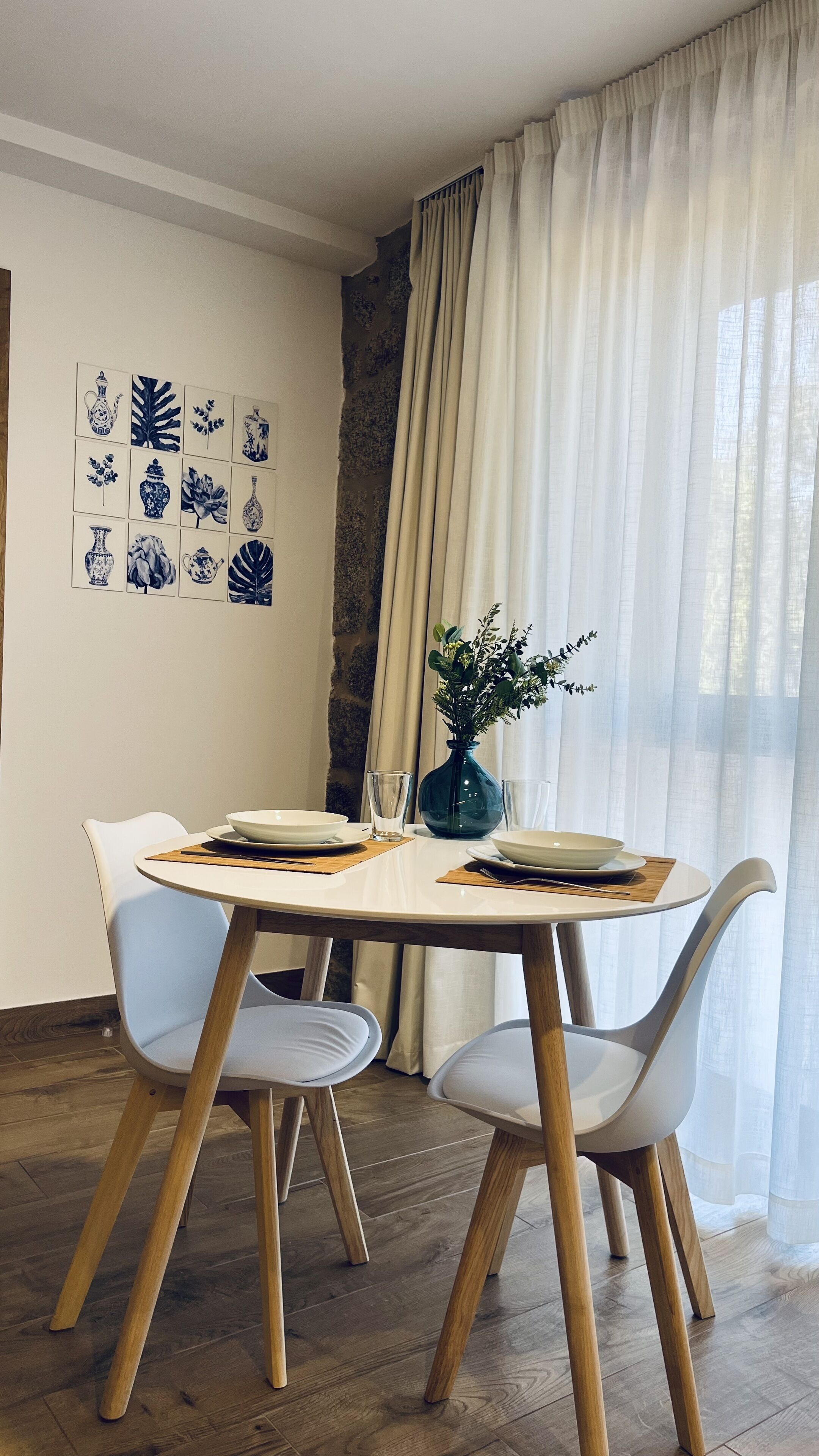 Un comedor con una mesa redonda, dos sillas, un florero con flores y decoración en la pared.