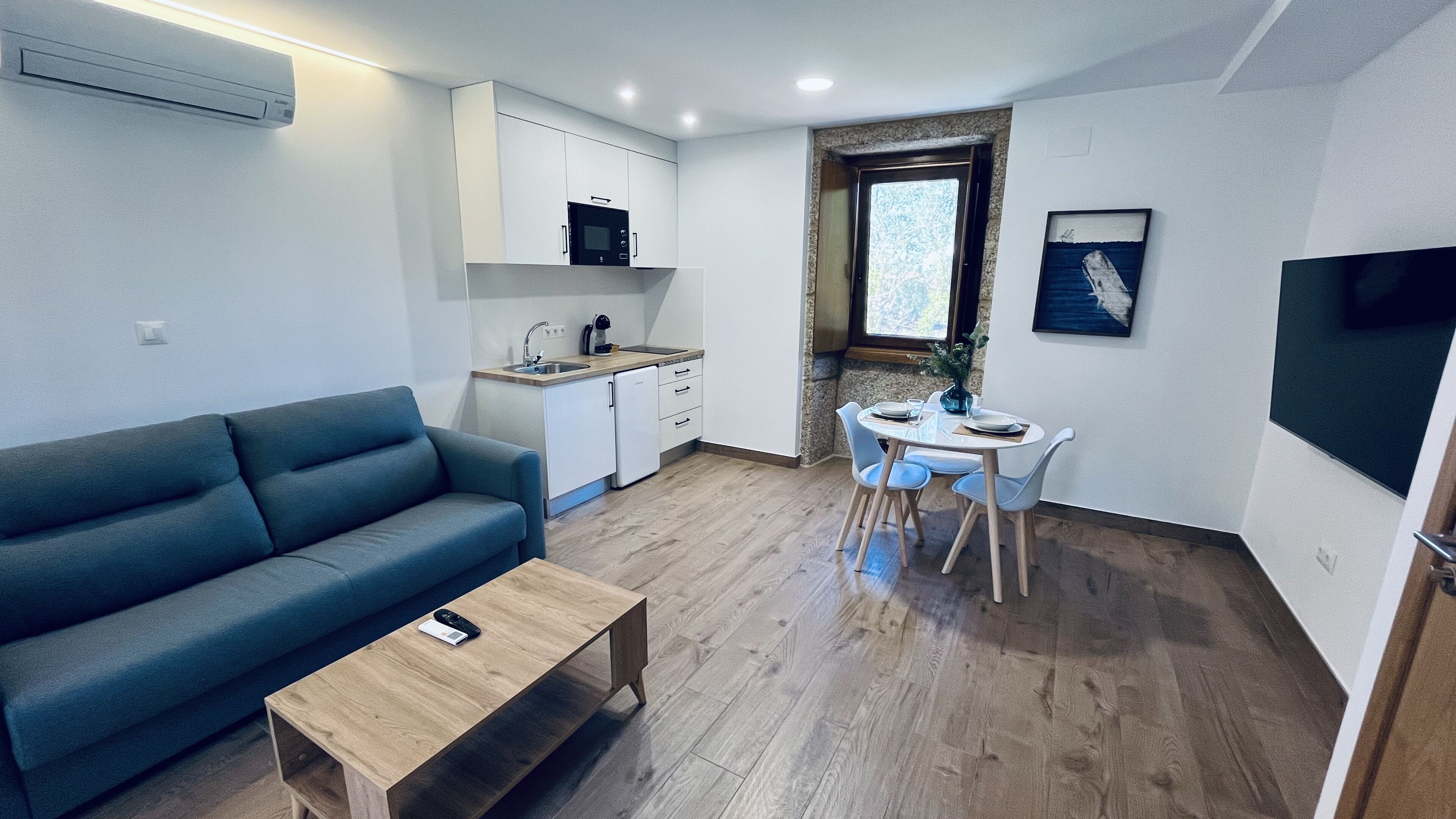 A compact living space with a sofa, a dining table with chairs, a kitchenette, and a TV mounted on the wall.