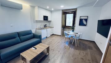 A compact living space with a sofa, a dining table with chairs, a kitchenette, and a TV mounted on the wall.
