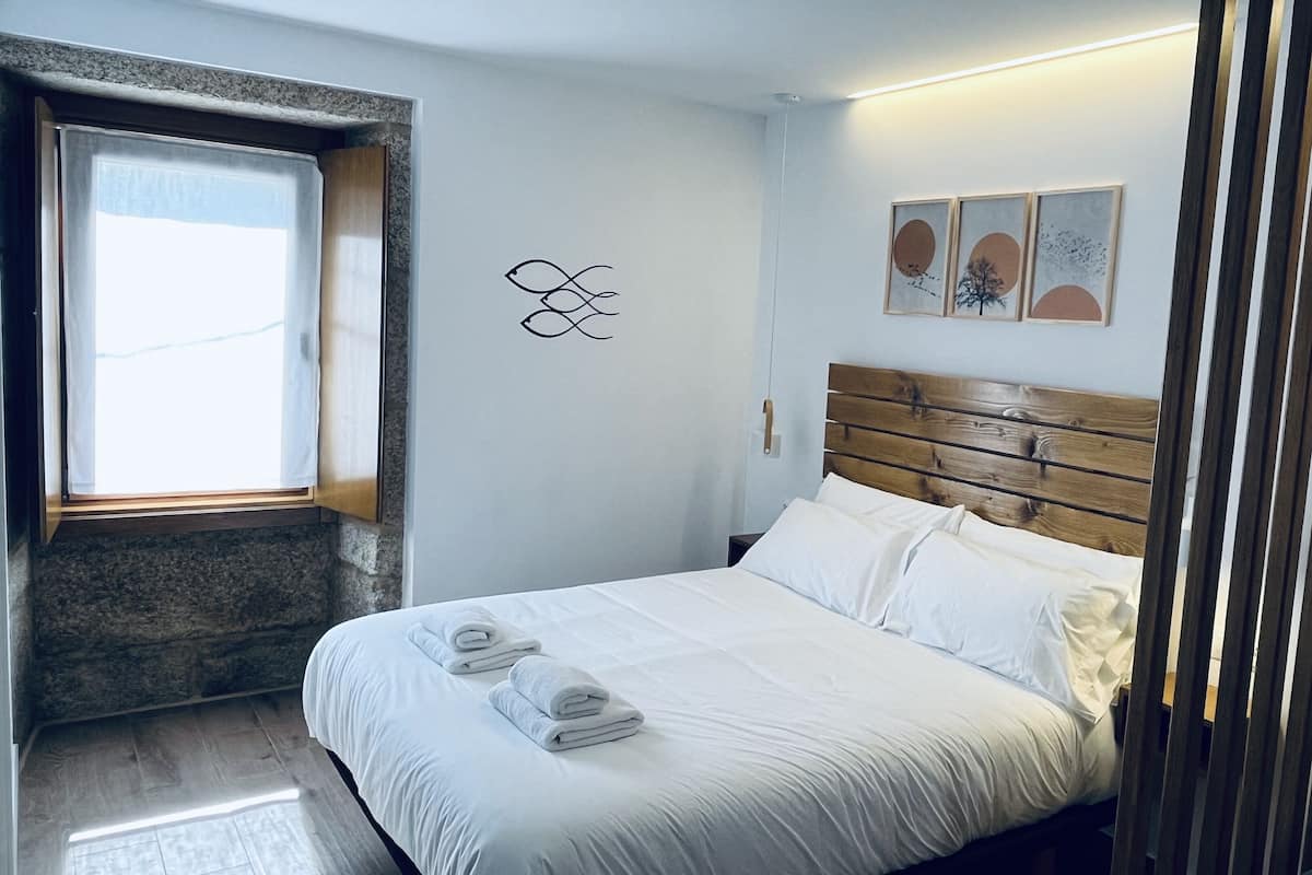 A neatly made bed with white linens and towels, a wooden headboard, and a window with curtains.