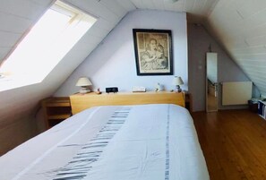 A bedroom with a sloped ceiling, a bed with a white bedspread, a nightstand with a lamp, a framed picture on the wall, and a radiator. - In the countryside, beaches just a few minutes away... (Plomeur)