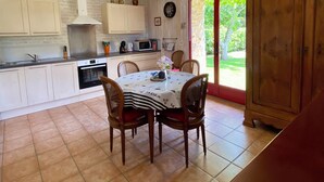 A kitchen with white cabinets, a dining table with chairs, a wooden cabinet, and a view of greenery outside. - In the countryside, beaches just a few minutes away... (Plomeur)
