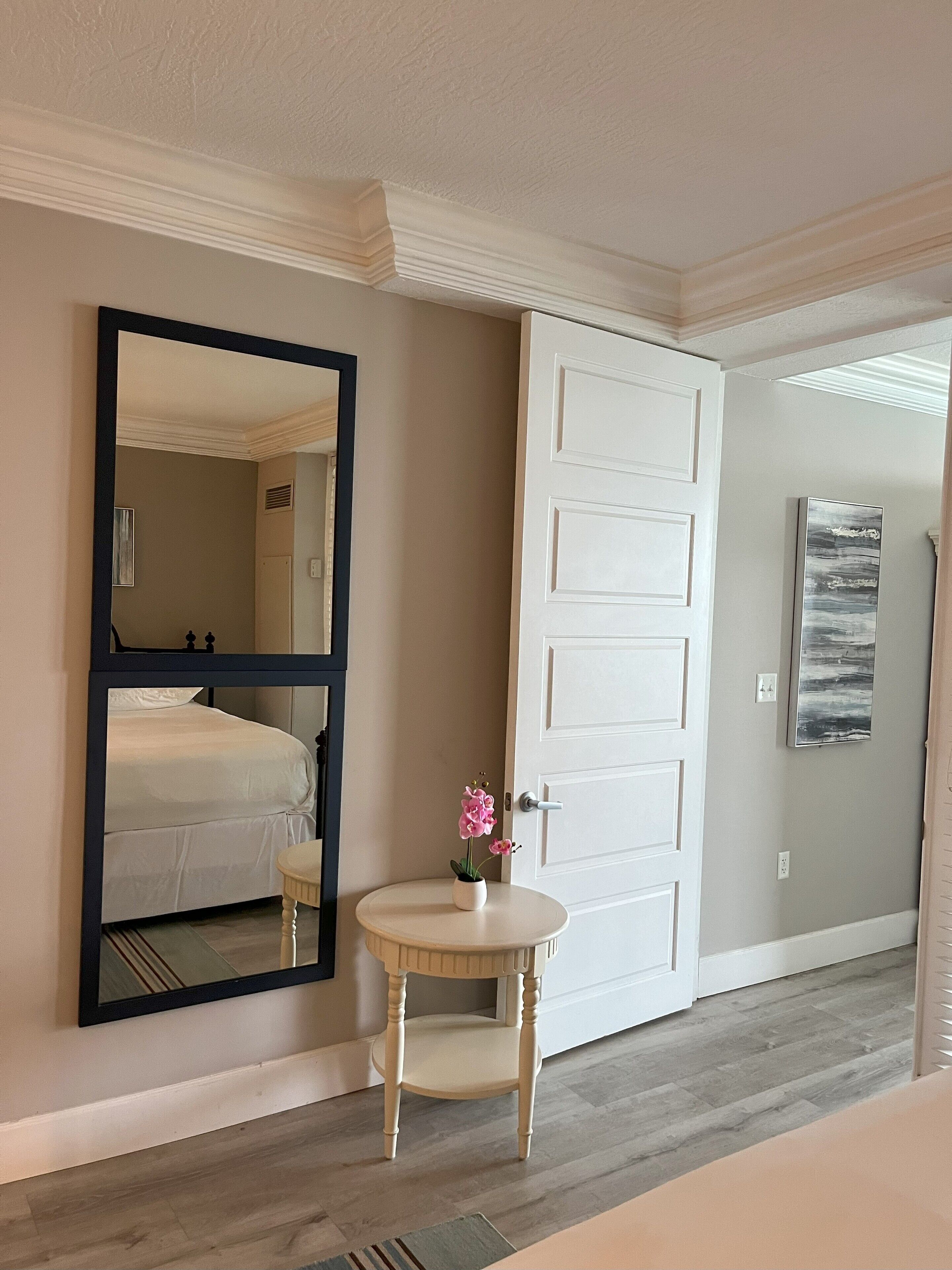 Une chambre d’hôtel avec un lit, une table ronde ornée d’un vase de fleurs, un grand miroir et une porte blanche.