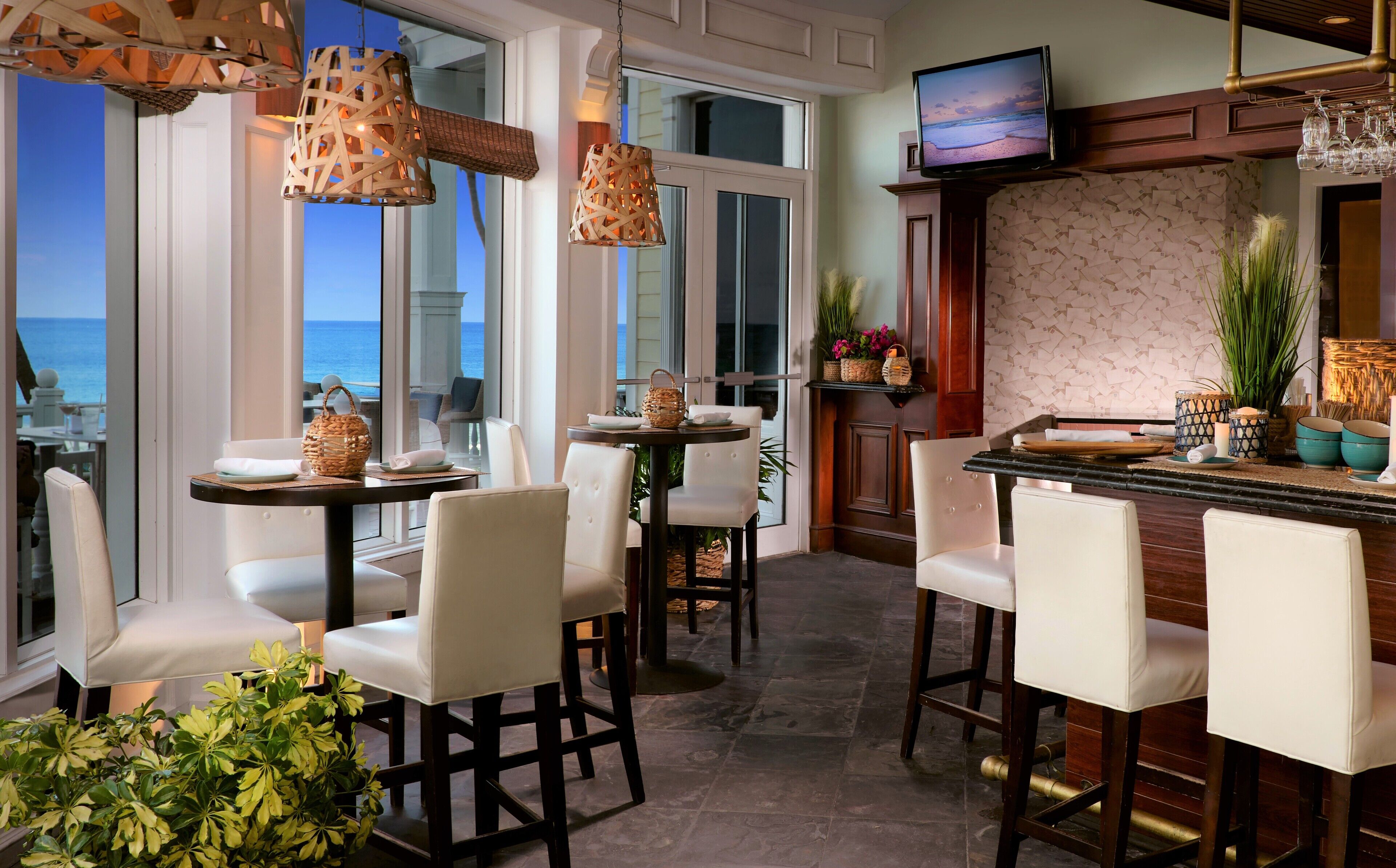 A bar area with white chairs, a television mounted above the bar, and a view of the ocean through large windows.