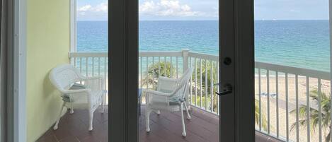 Un balcon avec une chaise blanche et une petite table, donnant sur une plage, l’océan et un ciel dégagé.