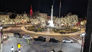Une vue nocturne d’une place publique avec une fontaine centrale, entourée d’arbres et illuminée par des lampadaires.