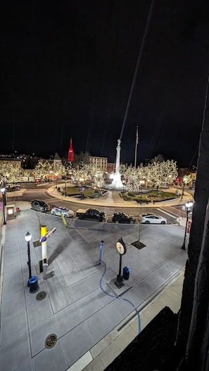 A nighttime view of a public square with a central fountain, surrounded by trees and illuminated by street lamps.