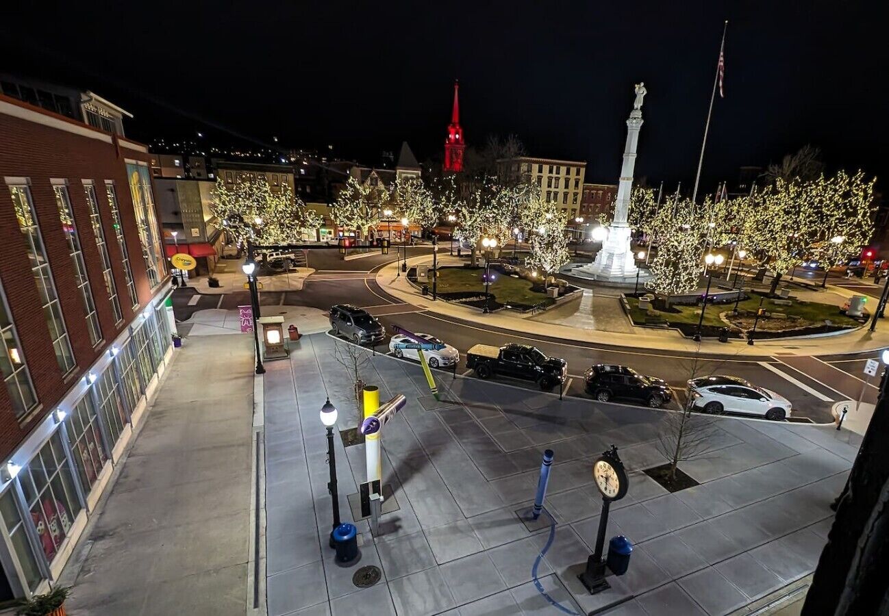 Une place urbaine illuminée la nuit, avec un monument central, une horloge et une tour rouge en arrière-plan.