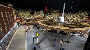Une place urbaine illuminée la nuit, avec un monument central, une horloge et une tour rouge en arrière-plan.