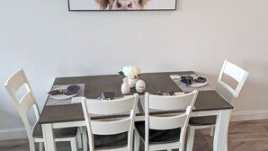 Ett matbord med fyra vita stolar, en inramad tavla med en ko på och en liten vit vas med en blomma.
