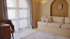 A living room with a sofa, a chair, and a table. There is a window with sheer curtains and a wall-mounted light fixture. - Malkata House - 32 ppl - Desert Oasis: Pool & Dine (Luxor)