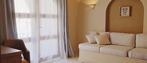 A living room with a sofa, a chair, and a table. There is a window with sheer curtains and a wall-mounted light fixture.