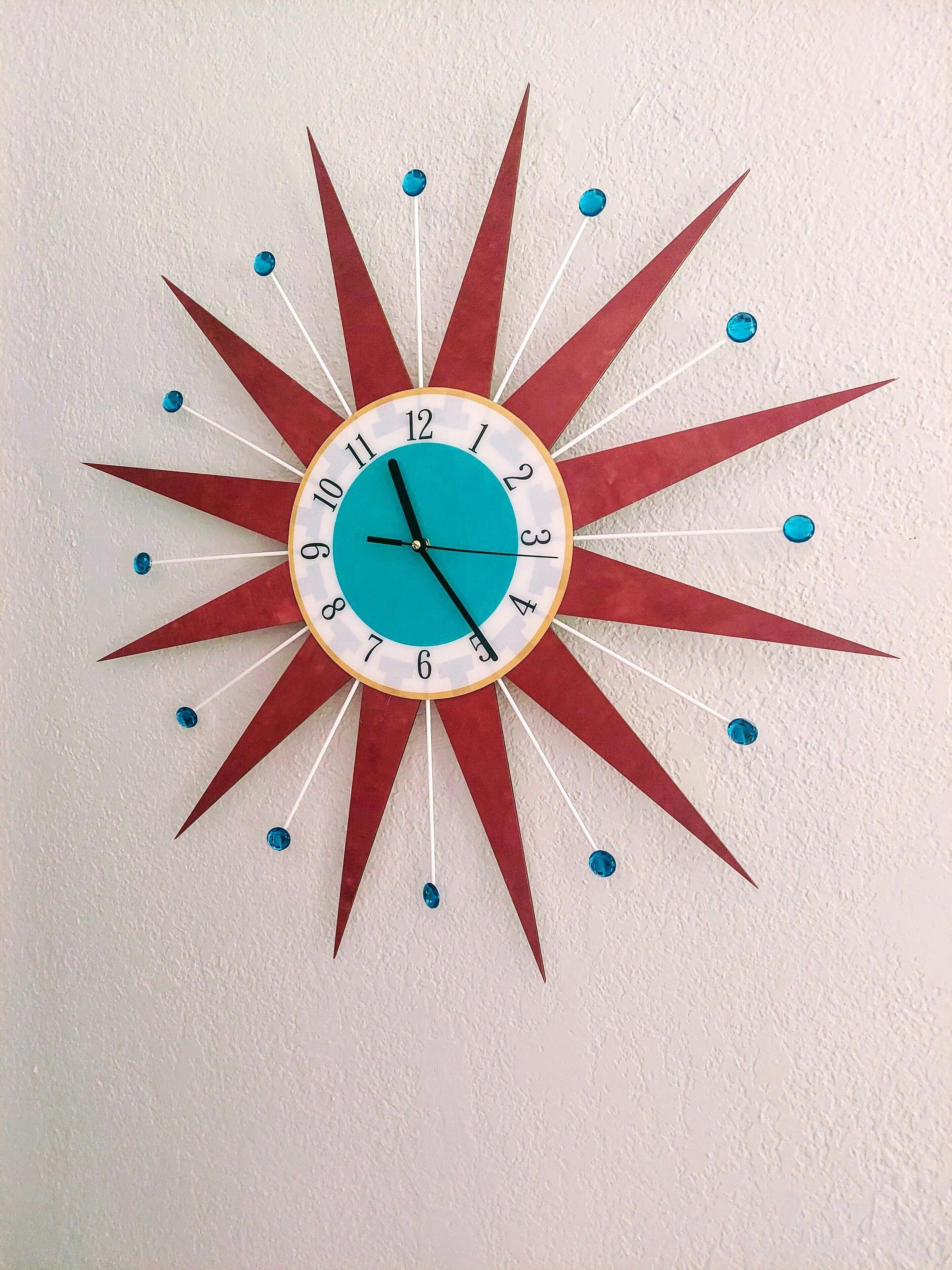 Een wandklok met een stralendesign, met een turquoise wijzerplaat en rode accenten.