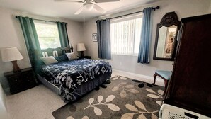 Un dormitorio con una cama, un ventilador de techo, una ventana con cortinas, un aparador y un espejo.
