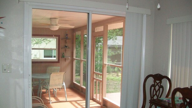 Una habitación con puerta de vidrio corredera que da a un balcón. En el interior, hay una mesa con sillas, un ventilador de techo y una ventana con persianas.