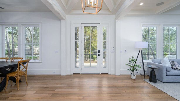 A spacious room with a wooden floor, white walls, and a large window. There is a dining table with chairs, a sofa, and a floor lamp. - Gorgeous new luxury beach house - be the first to experience this dream home! (Hilton Head)