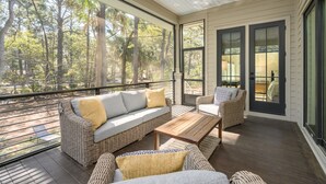 A covered outdoor seating area with wicker furniture, a wooden table, and large windows offering a view of a forested area. - Gorgeous new luxury beach house - be the first to experience this dream home! (Hilton Head)