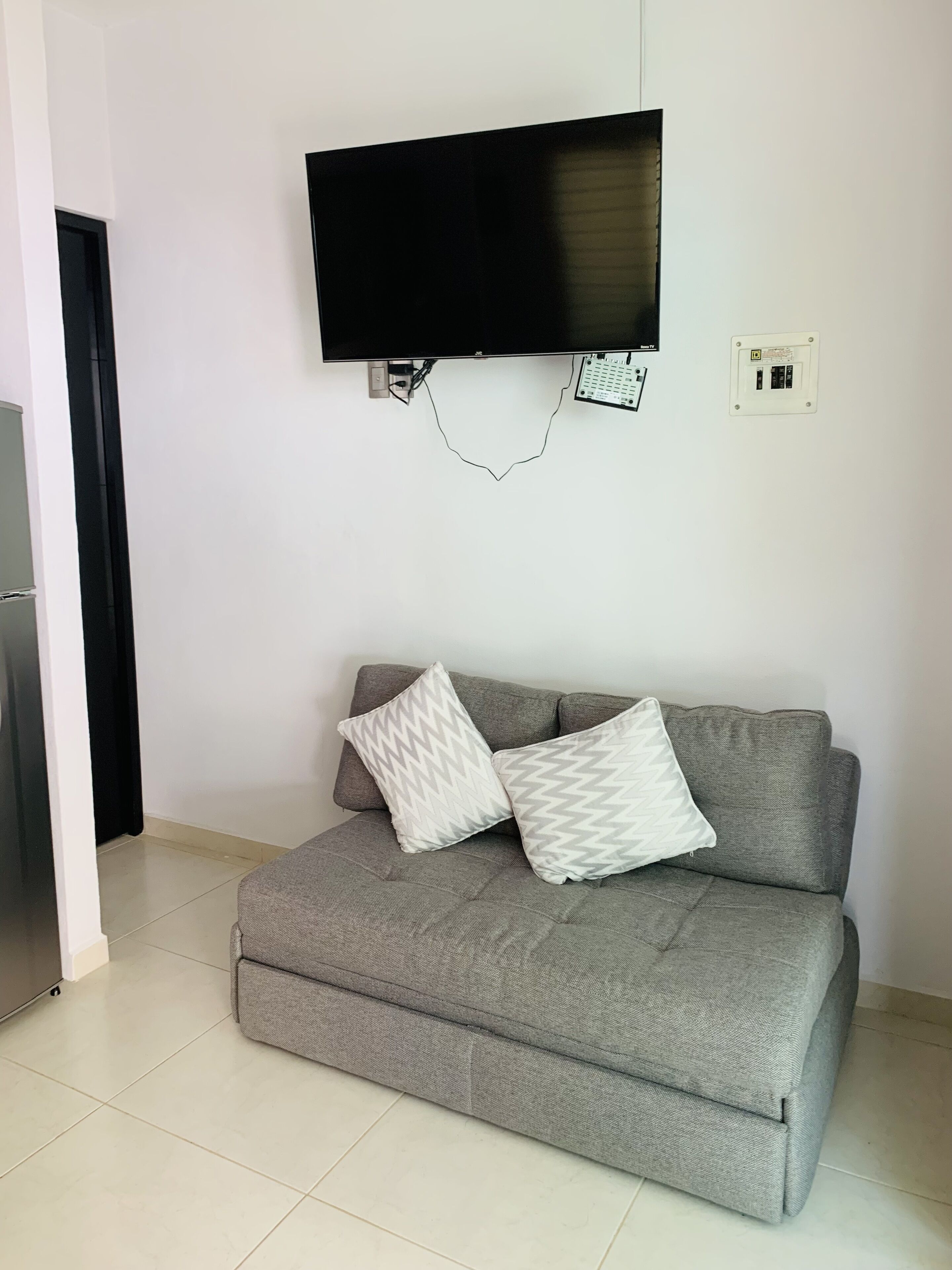 Ein modernes Wohnzimmer mit einem grauen Sofa, zwei gemusterten Kissen, einem an der Wand montierten Flachbildfernseher und einer Steckdose.
