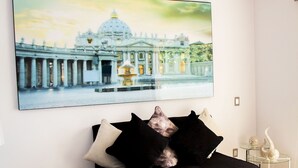 A modern living room with a black sofa, a large framed artwork of a building, and a glass side table with a decorative item.