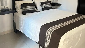 A modern bedroom with a black headboard, white bedding, and decorative pillows. There is a painting above the bed and a nightstand with a lamp on each side.