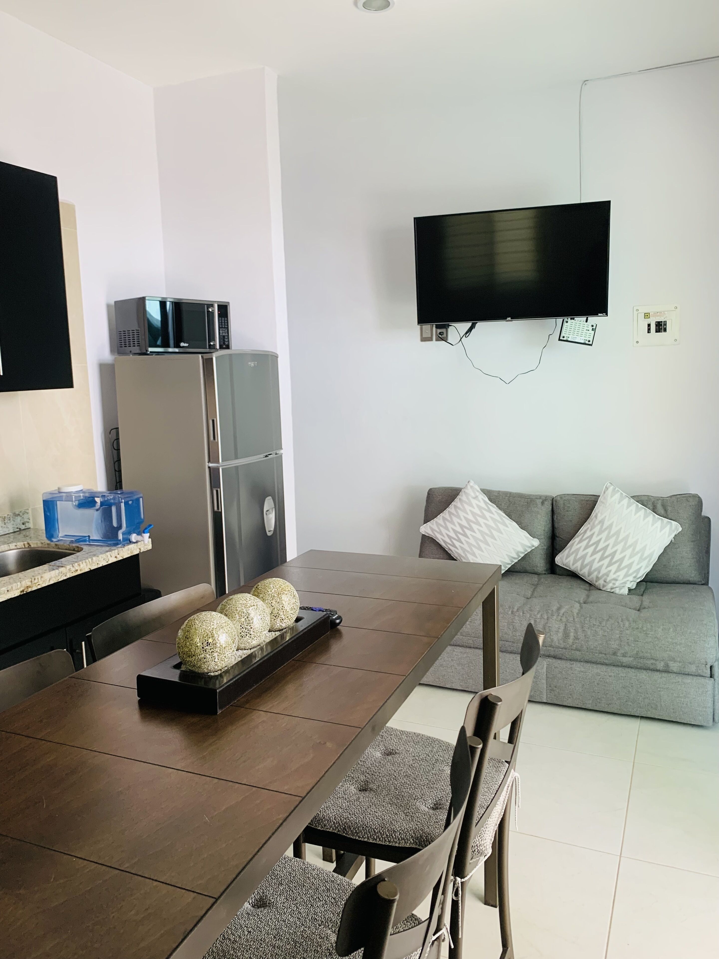 A modern kitchen with a built-in microwave, a refrigerator, and a water dispenser. There is a dining table with chairs and a flat-screen TV mounted on the wall.