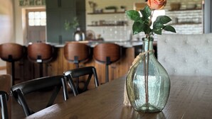 Een eetruimte met een houten tafel en stoelen, een vaas met een enkele roos, en een keuken op de achtergrond.