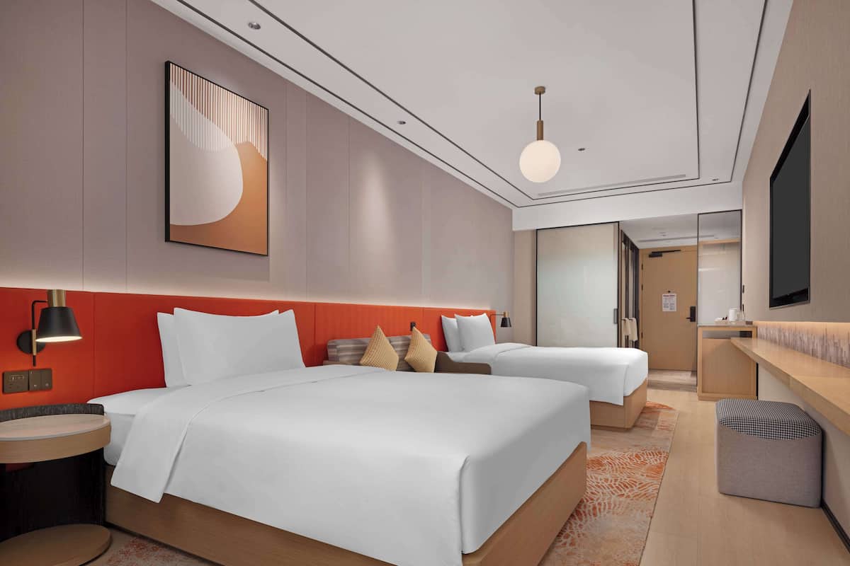 Une chambre d’hôtel moderne avec deux lits, une grande peinture abstraite, une petite table ronde, un petit pouf et un téléviseur fixé au mur.