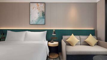 A hotel room with a bed, a sofa, a nightstand with a lamp, and a wall-mounted artwork.