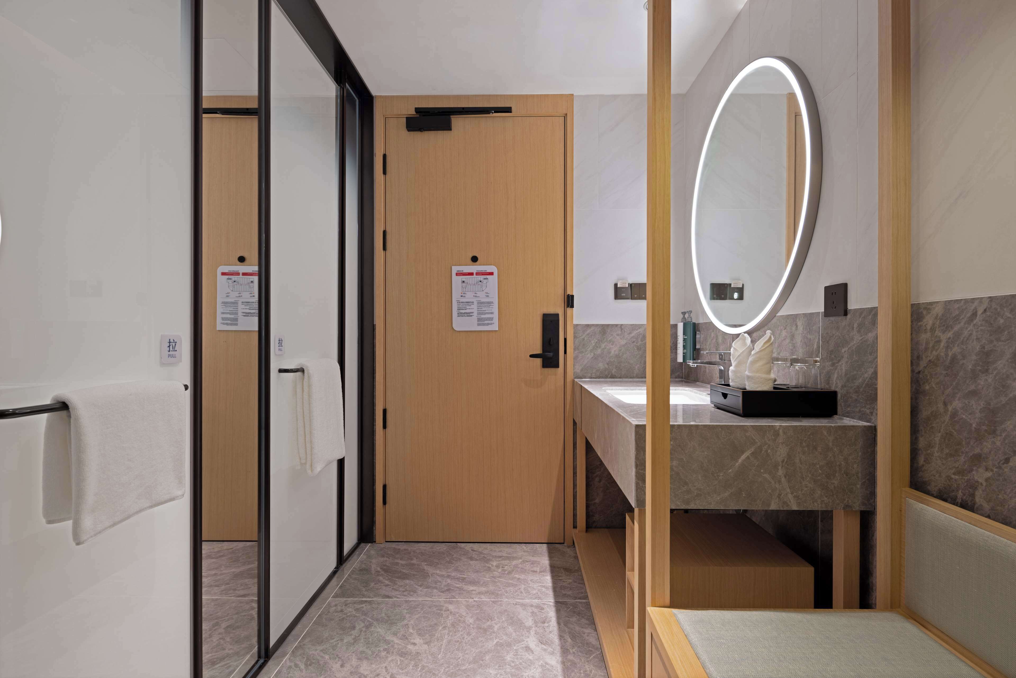 A modern bathroom with a wooden door, a mirror with a light, and a marble countertop.