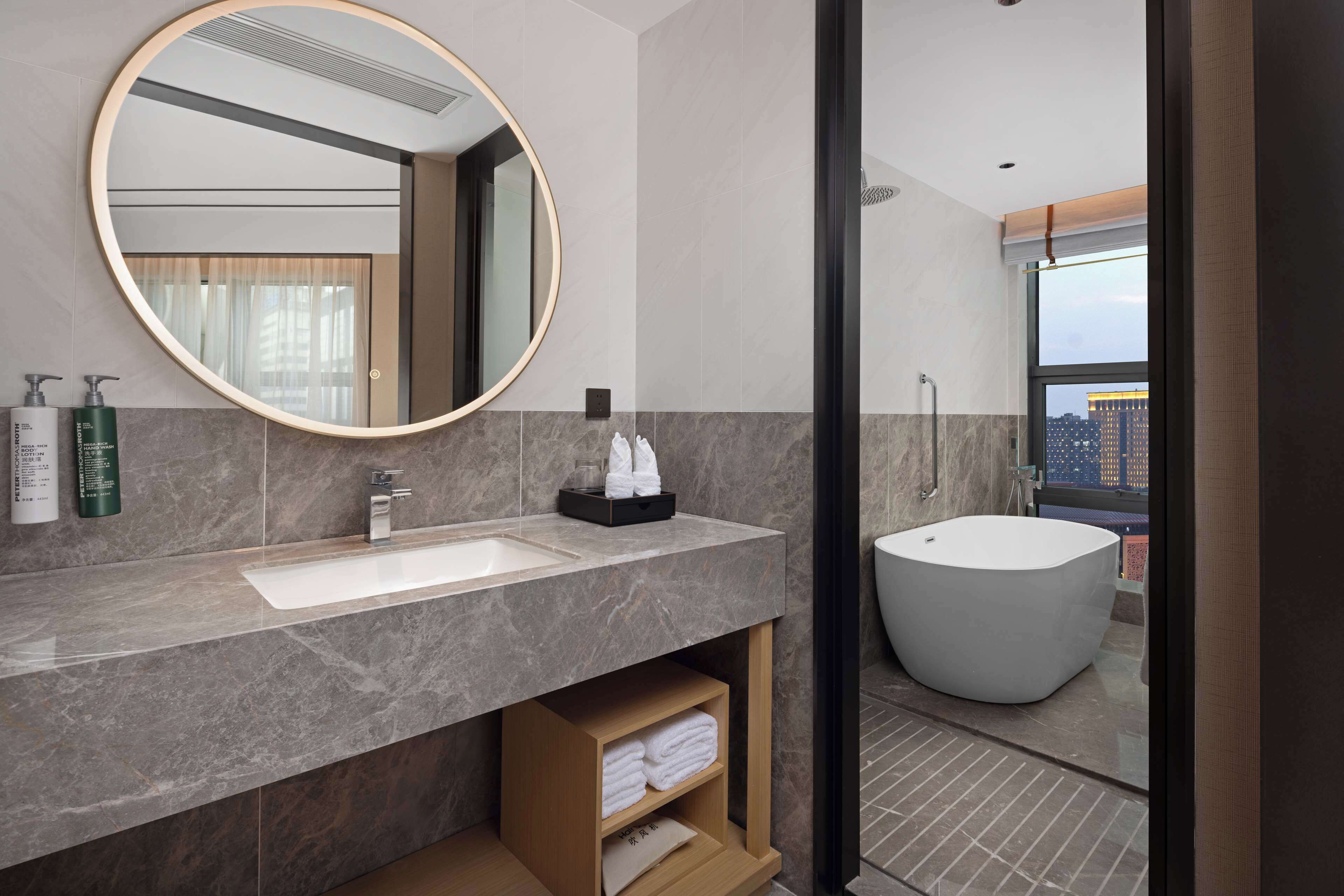 A modern bathroom with a large round mirror, a dual sink vanity, a bathtub, and a view of the city through a large window.