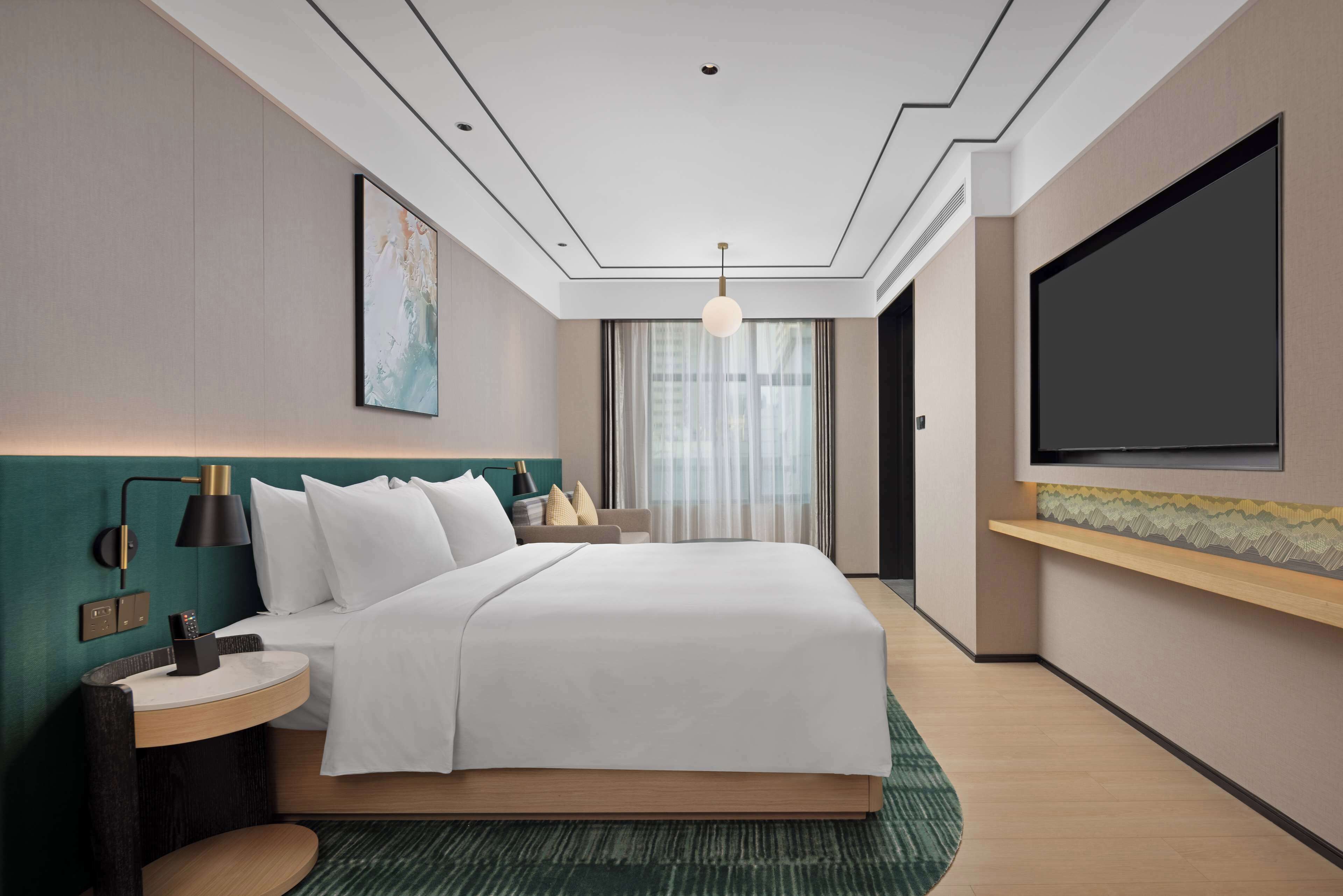 Ein modernes Hotelzimmer mit einem großen Bett, einem Nachttisch, einem an der Wand montierten Fernseher und einem großen Fenster mit Vorhängen.