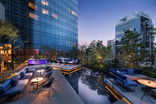 Un bar en plein air situé sur le toit d’un immeuble, avec des espaces pour s’asseoir et une piscine, le tout entouré de bâtiments modernes.