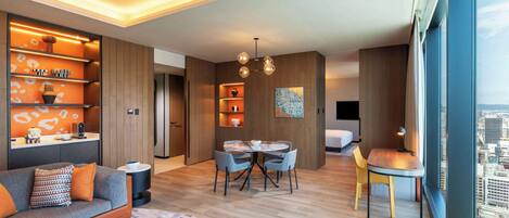 Habitación de hotel moderna con sofá, mesa de comedor, sillas, suelo de madera y vistas a la ciudad.