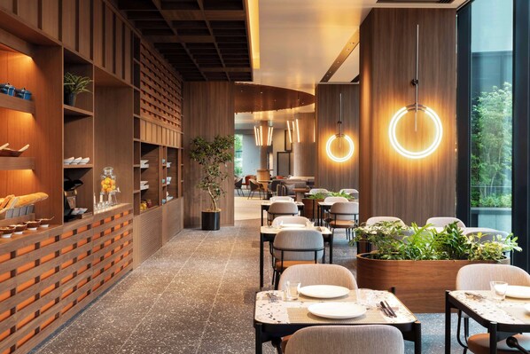Un restaurante moderno con paneles de madera, lámparas colgantes circulares y una exhibición central de plantas. Las mesas están preparadas con platos, cubiertos y copas.