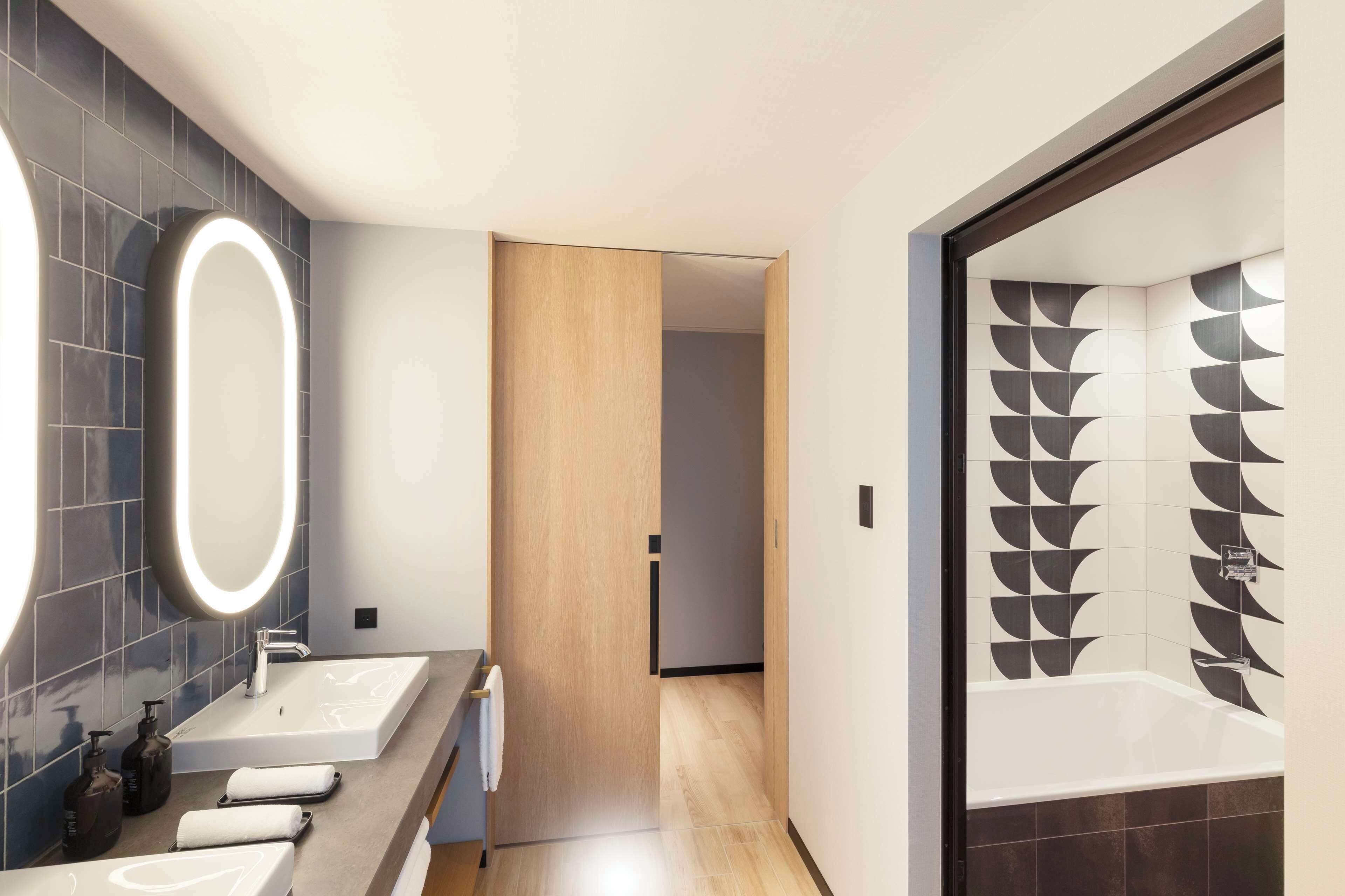 A modern bathroom with a large mirror, two sinks, and a bathtub with geometric tile design.