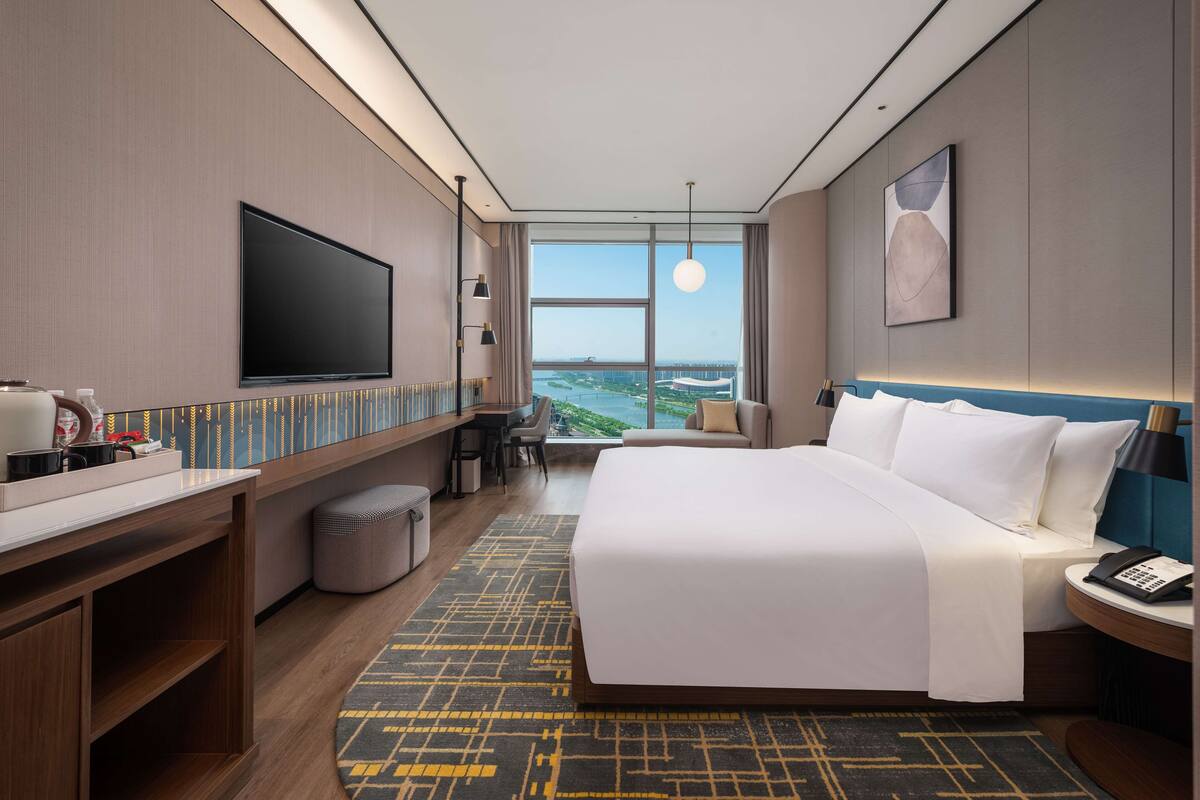 Une chambre d’hôtel moderne dotée d’un grand lit, d’un bureau avec un téléviseur à écran plat, d’un coin salon offrant une vue sur un pont et d’une grande fenêtre.
