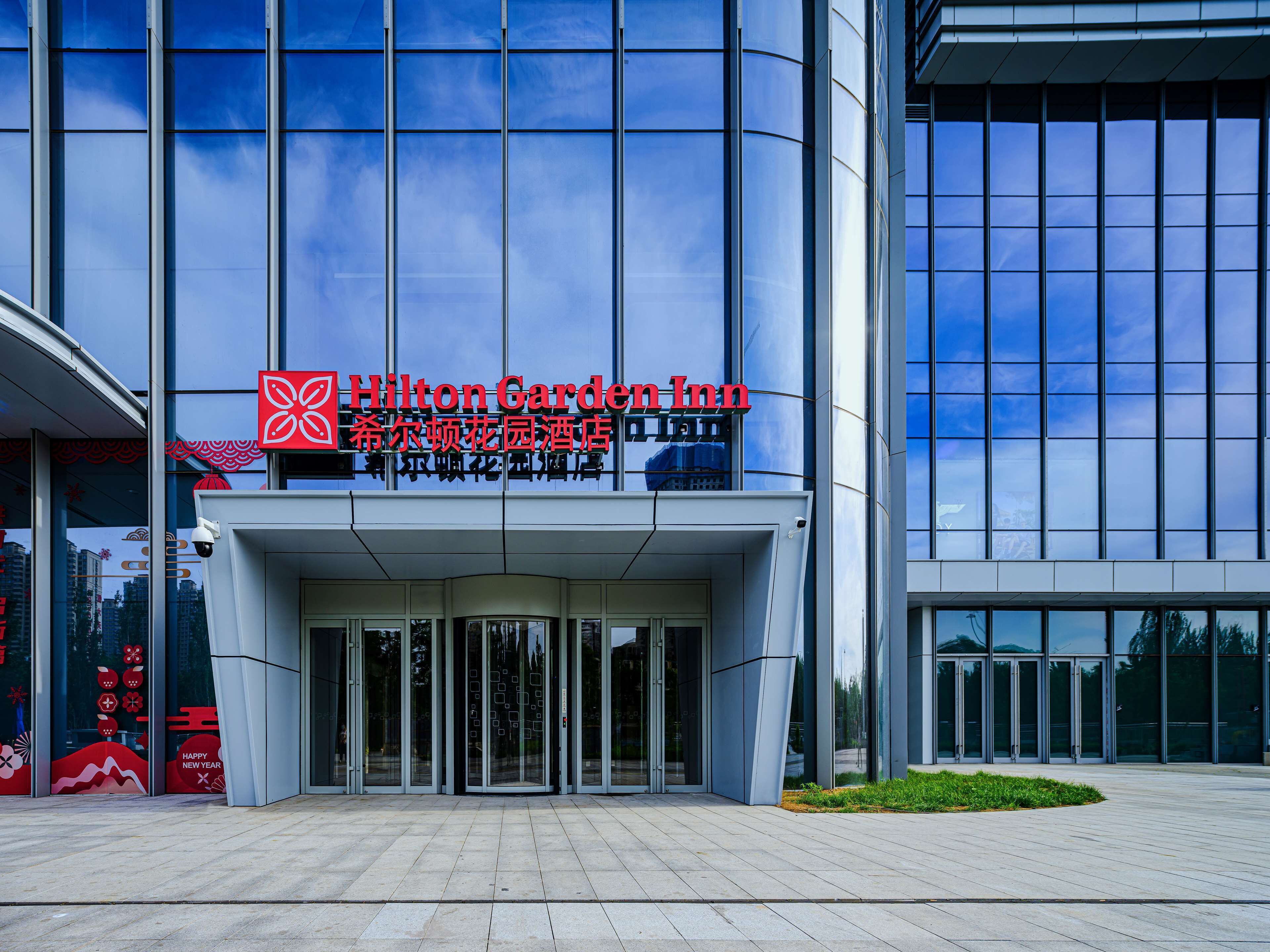 The exterior of a Hilton Garden Inn with a modern glass facade, large entrance doors, and a clear sky in the background.