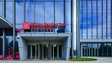 The exterior of a Hilton Garden Inn with a modern glass facade, large entrance doors, and a clear sky in the background.
