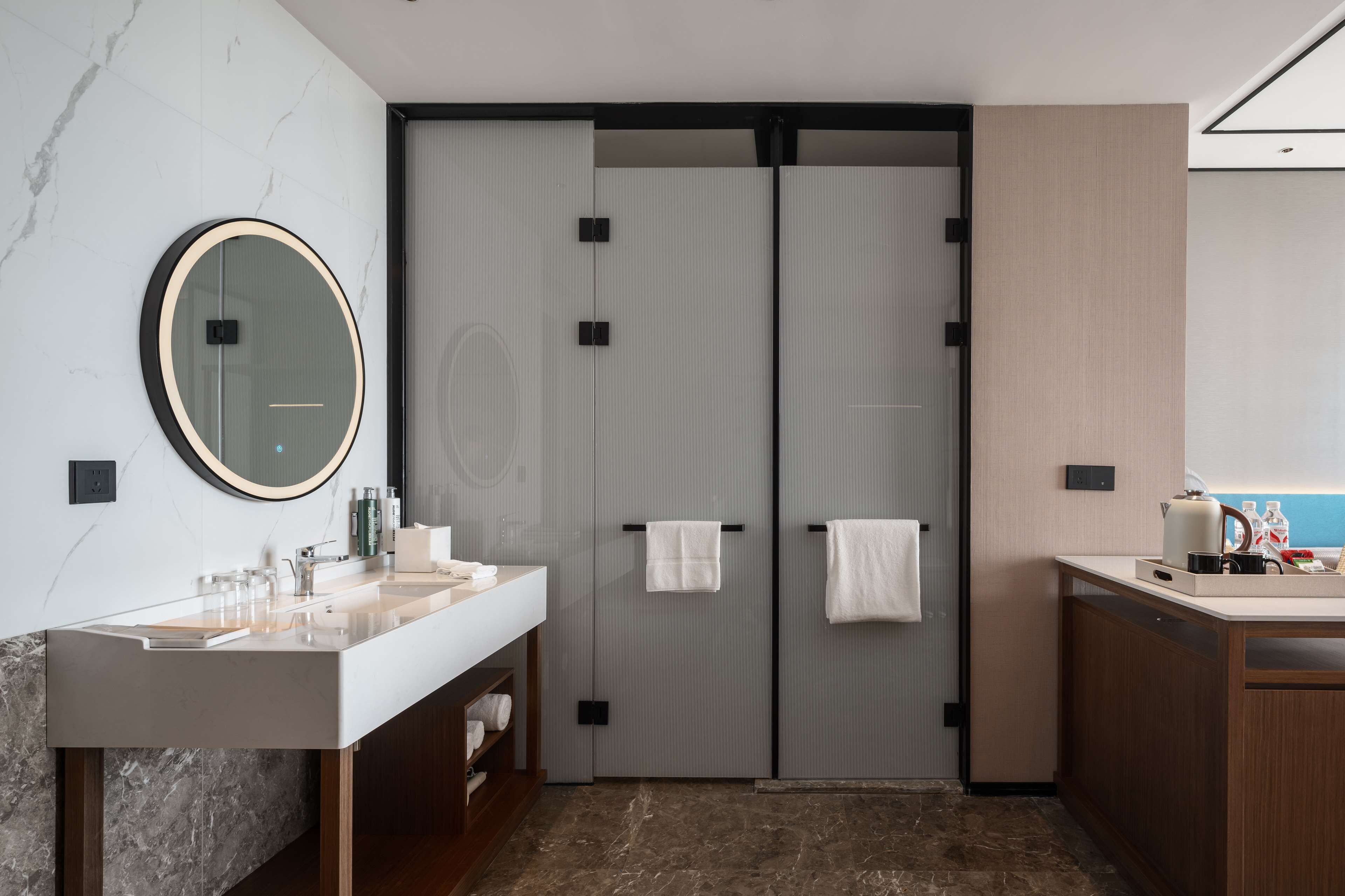 Un baño moderno con un espejo grande, dos barras para toallas, un lavabo con artículos de aseo y una pequeña cocina con hervidor.