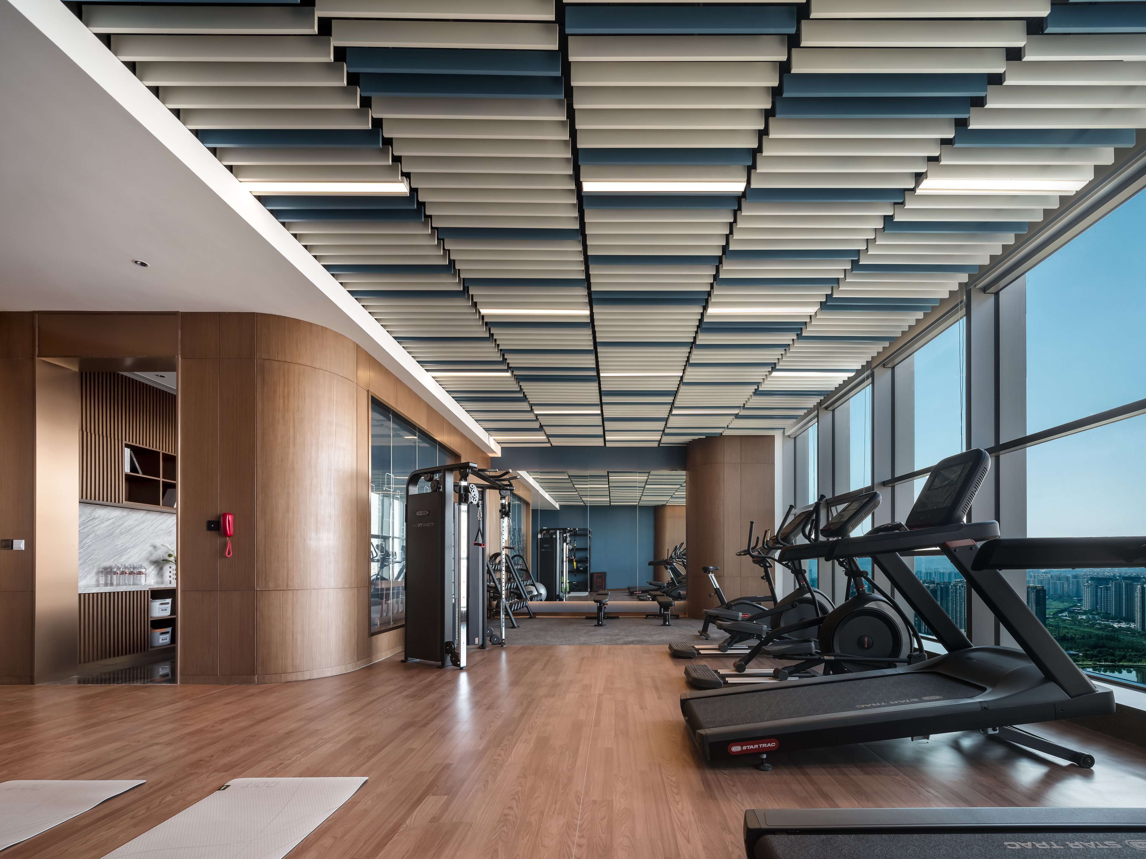A modern gym with wooden floors, exercise equipment, and large windows offering a city view.