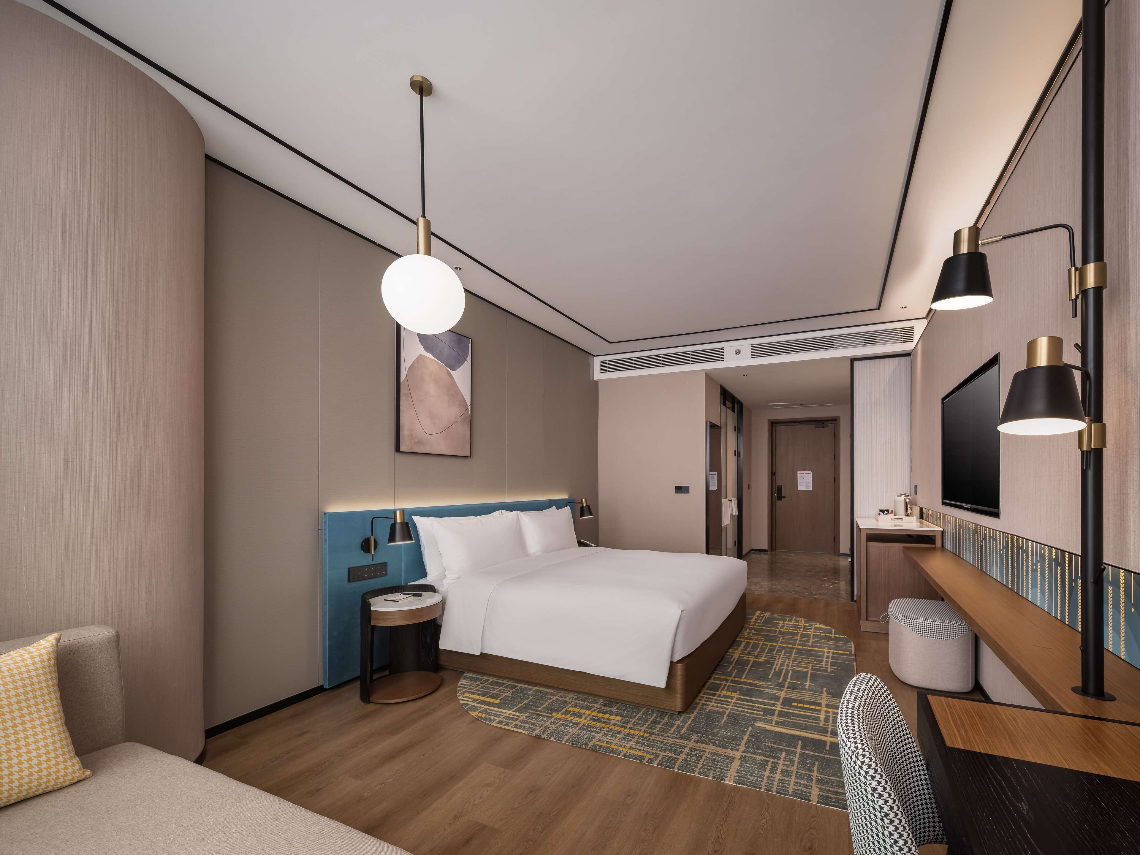 A modern hotel room with a large bed, bedside tables, a desk with a chair, a TV, and a large mirror.