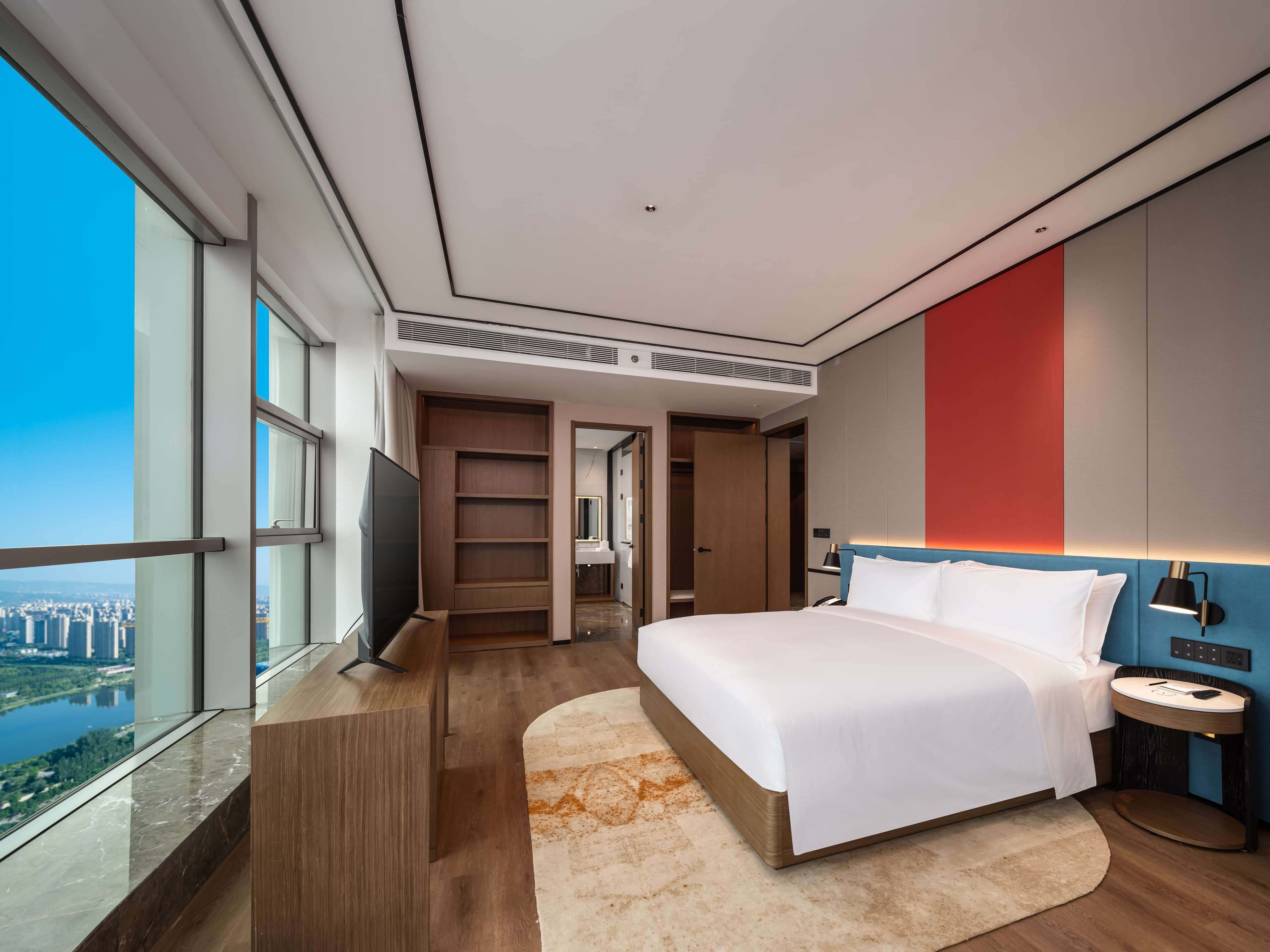 Ein modernes Hotelzimmer mit einem großen Bett, einem Fernseher, einem begehbaren Kleiderschrank und Blick auf die Stadt.