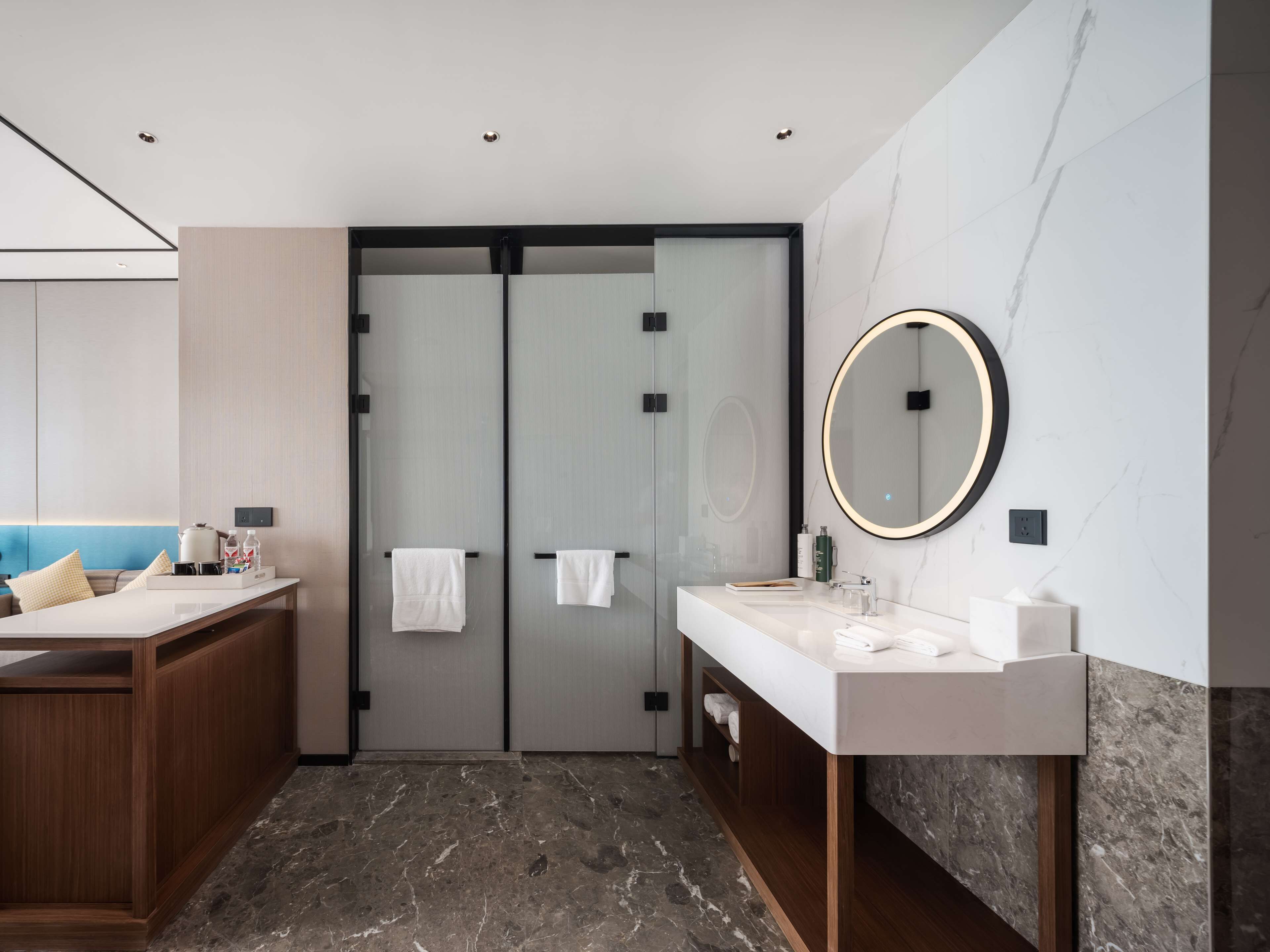 A modern bathroom with a marble floor, a wooden vanity, a round mirror, and a double door with towels.
