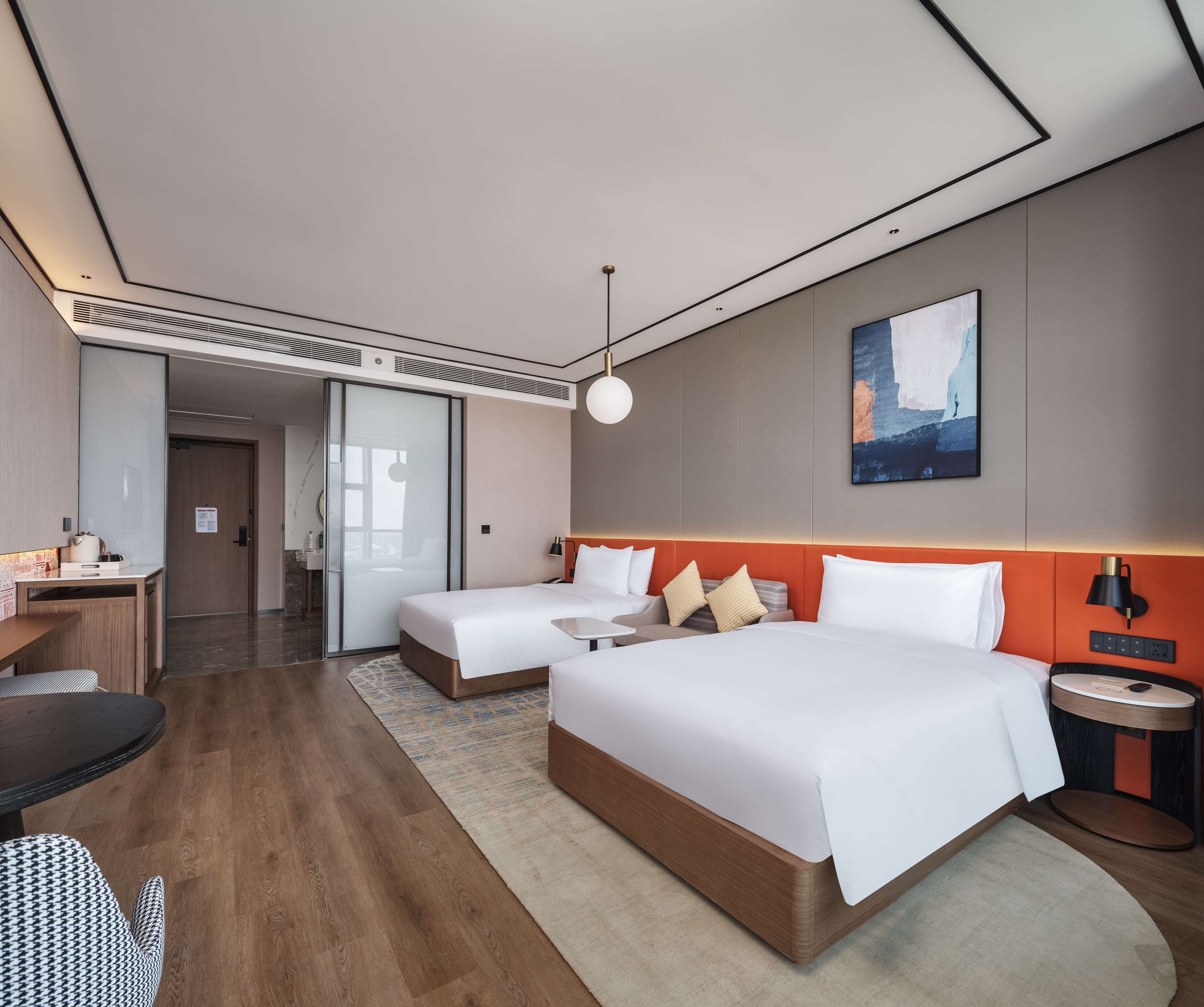 Ein modernes Hotelzimmer mit zwei Betten, einem Schreibtisch, einem Stuhl, einem kleinen Tisch und einer Wandlampe.