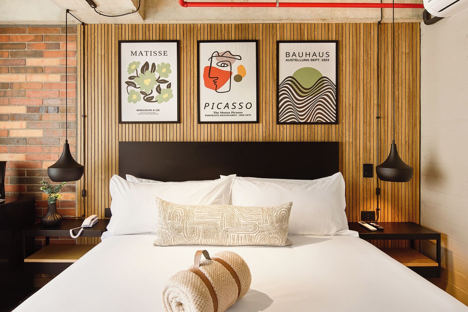 A hotel room with a bed, a nightstand, a lamp, and three framed artworks on the wall.
