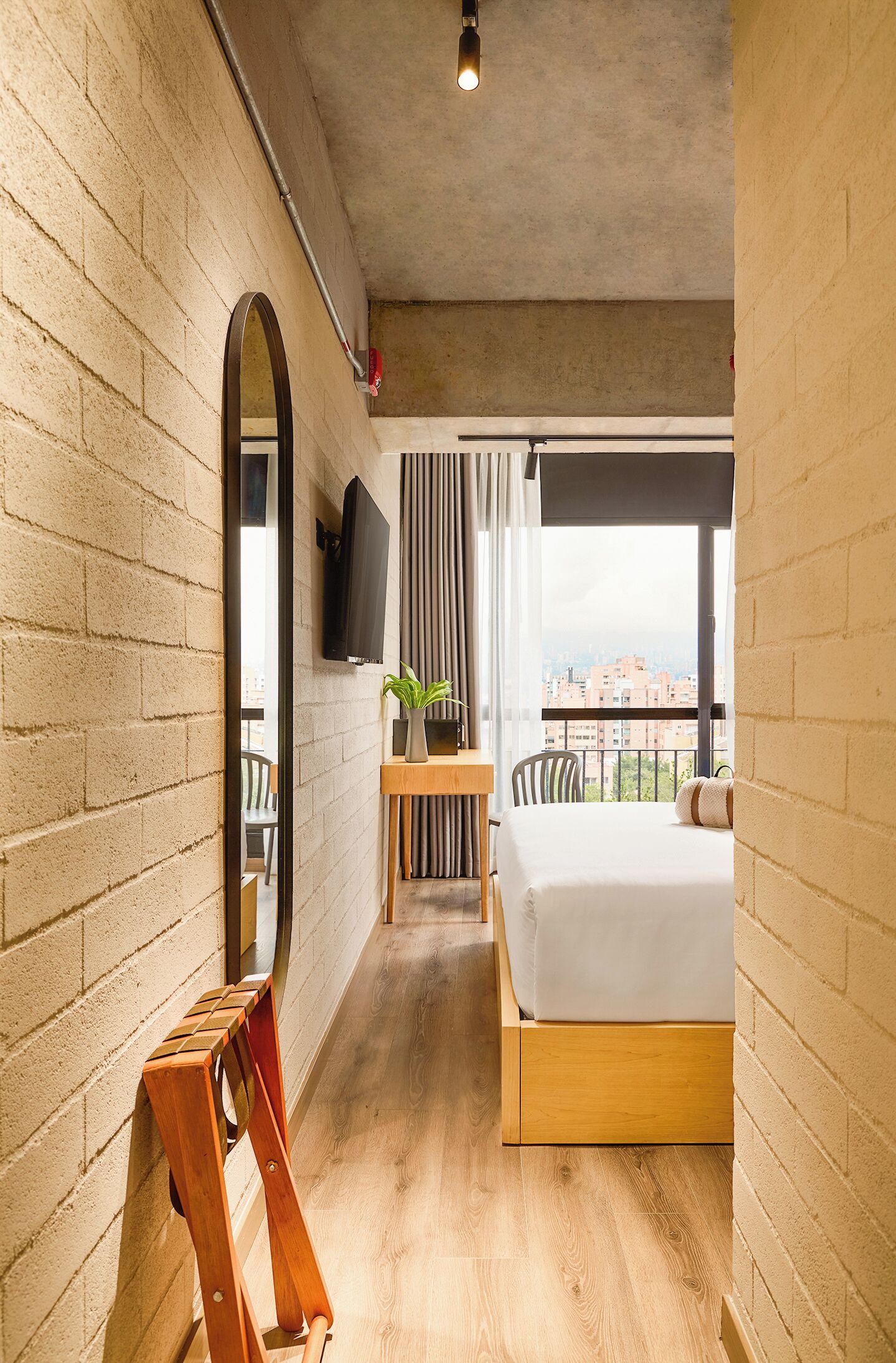 A bedroom with a bed, a wooden desk, a chair, a TV, and a view of the city.