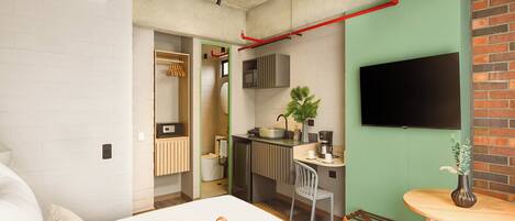 Habitación de hotel pequeña y funcional con cama, kitchenette, televisor montado en la pared y baño con ducha e inodoro.
