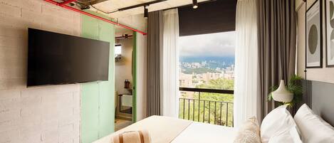 A modern hotel room with a large bed, a flat-screen TV mounted on the wall, and a balcony with a city view.