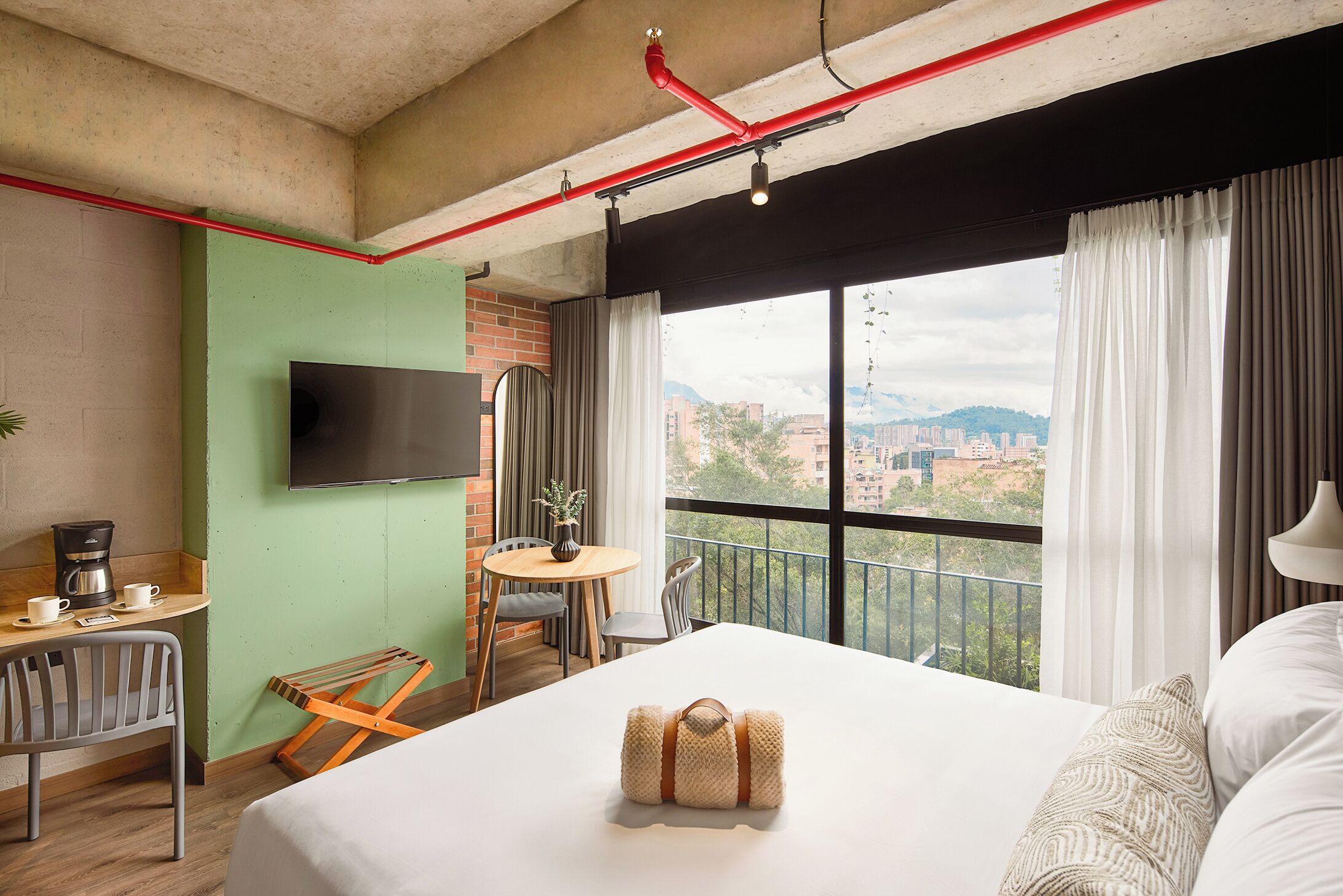 A hotel room with a bed, a desk with a coffee maker, a TV, a balcony with a view of the city, and a window with curtains.